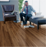 Man sitting on a couch with a laptop and papers, talking on a phone, in a room with wooden flooring.